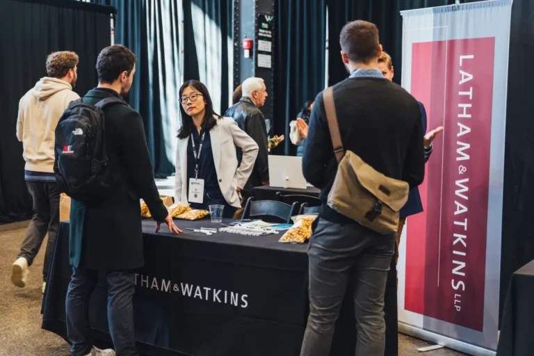 People interacting at a booth with Latham & Watkins branding, snacks on the table, in an expo hall setting.