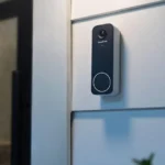 Black video doorbell mounted on a white exterior wall, with a window showing a lit lamp inside.