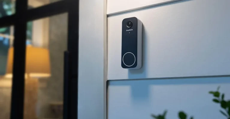 Black video doorbell mounted on a white exterior wall, with a window showing a lit lamp inside.