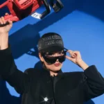 Person wearing a dark coat, cap with text, and sunglasses holds a red chainsaw against a blue background.