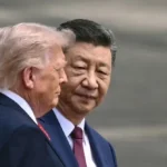 Two men in suits walking closely together, one with light hair and the other with dark hair, in an outdoor setting.