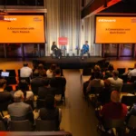 Audience watching two people on stage at a StrictlyVC event, discussing with Mark Rostick, with screens displaying event d...