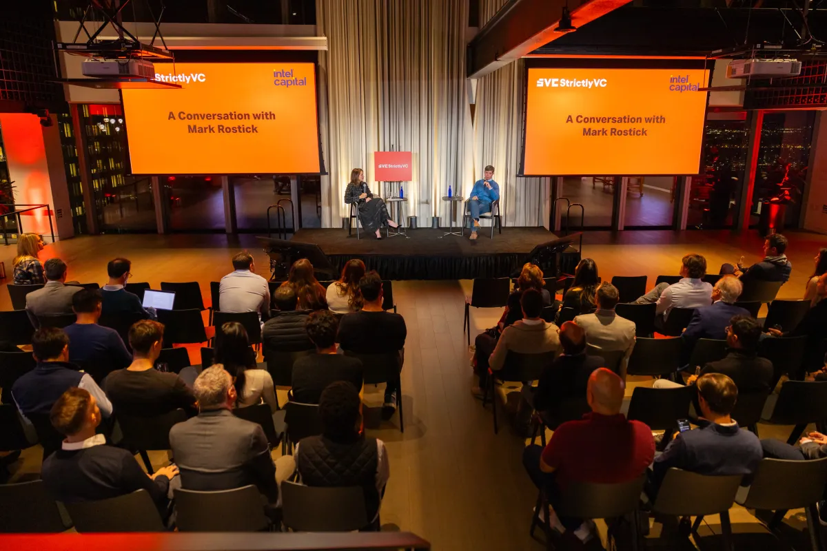 Audience watching two people on stage at a StrictlyVC event, discussing with Mark Rostick, with screens displaying event d...