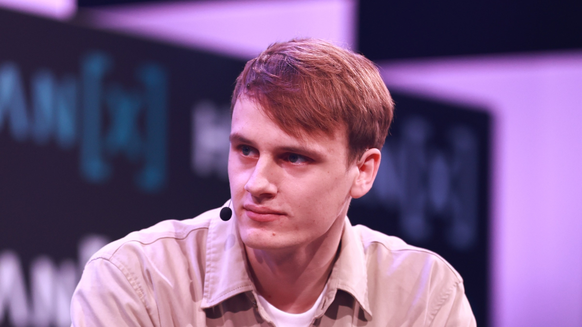 Person with a microphone headset, wearing a beige shirt, sitting on stage with blurred background featuring text and graph...