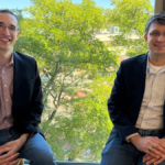 Two men in blazers sitting on a windowsill, smiling, with a leafy tree visible outside the window.