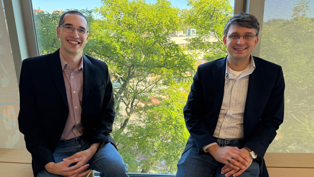Two men in blazers sitting on a windowsill, smiling, with a leafy tree visible outside the window.