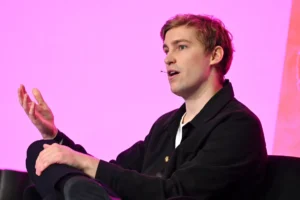 Person speaking with a headset, gesturing with hand, against a pink background.
