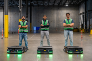 Three workers in green vests stand with arms crossed behind autonomous robots labeled 1, 2, and 3 in a warehouse.