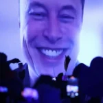 Large smiling face projected on screen, crowd in foreground holding up phones and cameras, blue and purple lighting