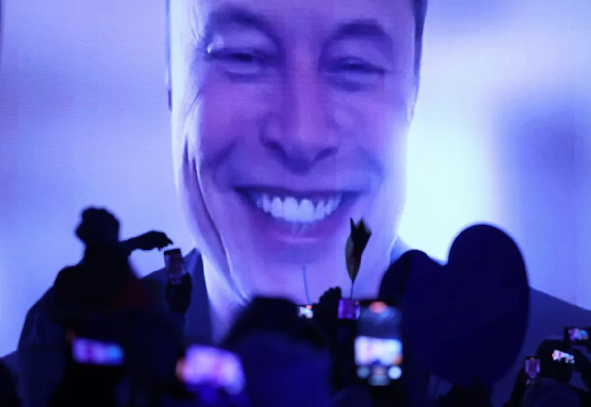 Large smiling face projected on screen, crowd in foreground holding up phones and cameras, blue and purple lighting