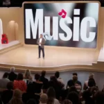 Presenter on stage with large "Music" display, audience seated, red speakers and "On Air" sign visible.