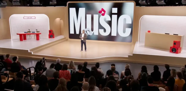 Presenter on stage with large "Music" display, audience seated, red speakers and "On Air" sign visible.