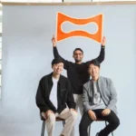 Three people pose indoors; two sitting, one standing holding an orange logo above them against a light gray backdrop.