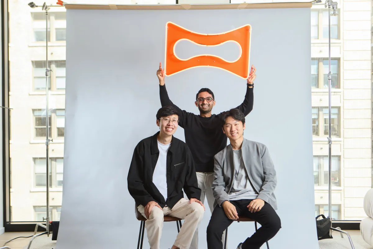 Three people pose indoors; two sitting, one standing holding an orange logo above them against a light gray backdrop.