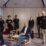Seven people in an office setting, five wearing black shirts with "Apate" logos, one seated in a chair, others standing.