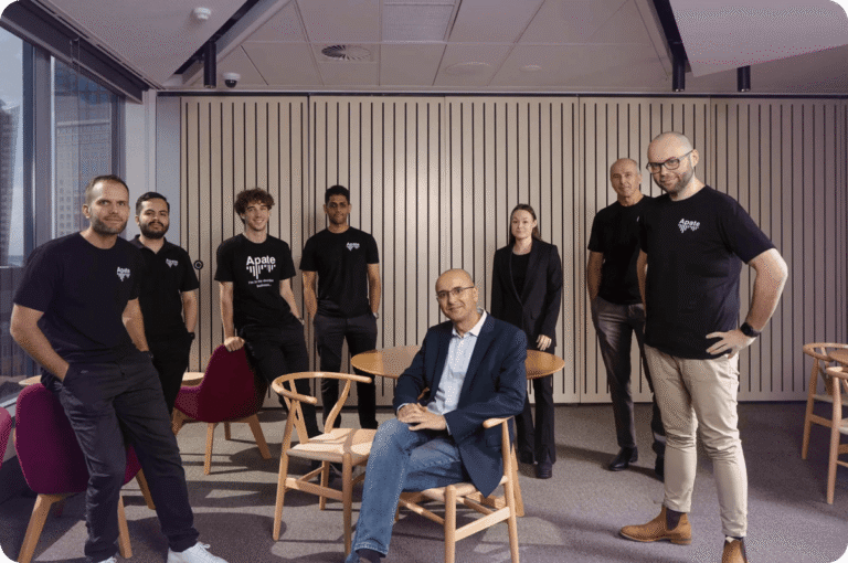 Seven people in an office setting, five wearing black shirts with "Apate" logos, one seated in a chair, others standing.