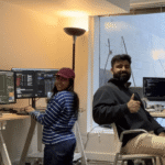 Two people smiling at computer workstations, one giving a thumbs-up, multiple screens displaying code and data.