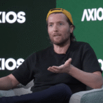 Man in a yellow cap speaking on stage with Axios logos in the background, gesturing with one hand.