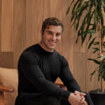 Man in black sweater seated on a wooden chair in a room with wood panel walls and a large potted plant.