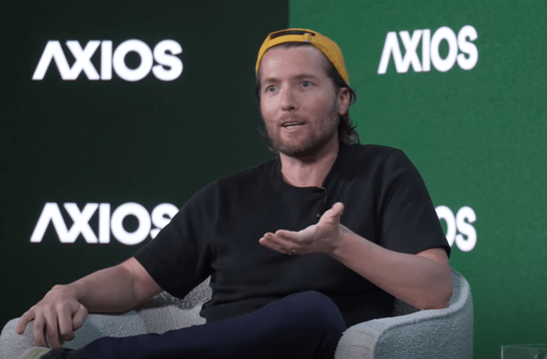 Man in a yellow cap speaking on stage with Axios logos in the background, gesturing with one hand.