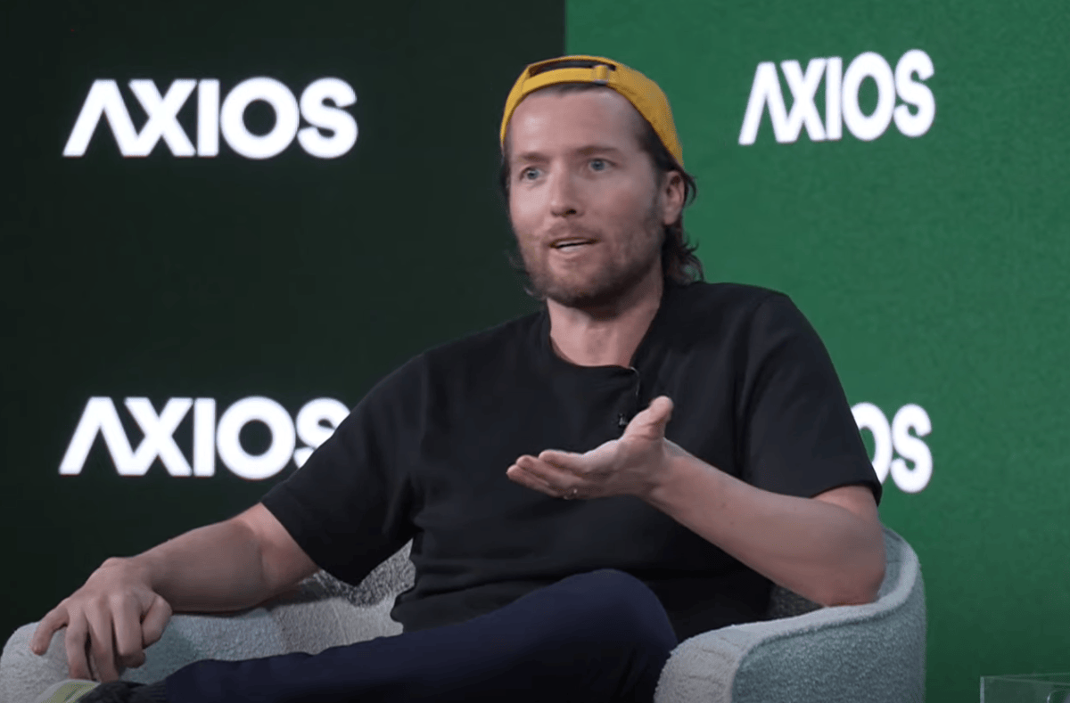 Man in a yellow cap speaking on stage with Axios logos in the background, gesturing with one hand.