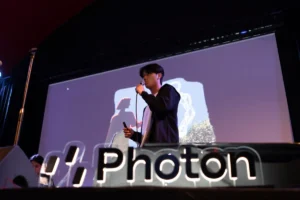 Person speaking on stage with microphone, large "Photon" sign in foreground, abstract graphics projected behind.