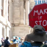 Person in a crowd holding a red octagonal sign with "MAKE it STOP" in white letters, urban setting in background.