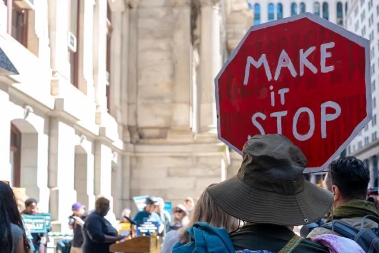 Person in a crowd holding a red octagonal sign with "MAKE it STOP" in white letters, urban setting in background.