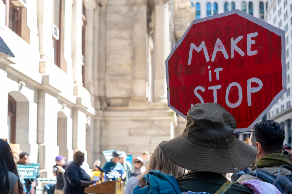 Person in a crowd holding a red octagonal sign with "MAKE it STOP" in white letters, urban setting in background.