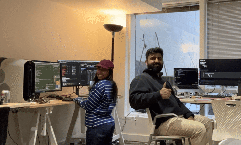 Two people smiling at computer workstations, one giving a thumbs-up, multiple screens displaying code and data.