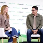 Two people seated and talking on stage at TechCrunch Disrupt, backdrop with event logos.