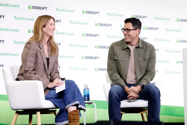 Two people seated and talking on stage at TechCrunch Disrupt, backdrop with event logos.