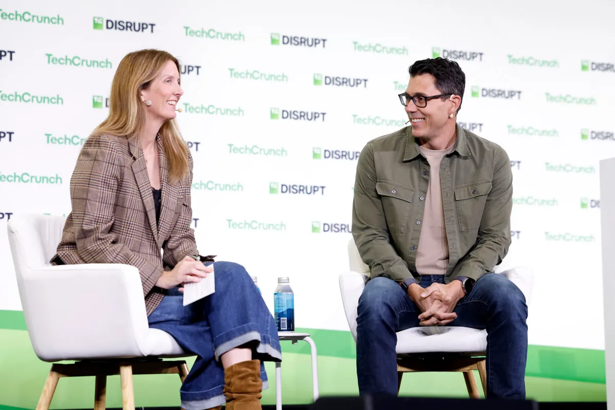 Two people seated and talking on stage at TechCrunch Disrupt, backdrop with event logos.
