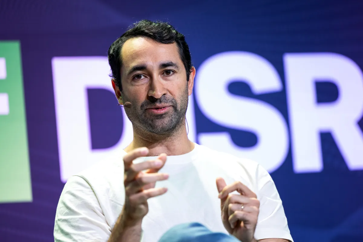 Man speaking on stage with a headset mic, gesturing with hands, large partial letters "DSR" visible in the background.
