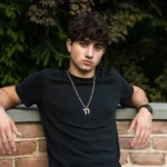 Person in a black shirt leans against a brick wall with greenery in the background, wearing a necklace and watch.