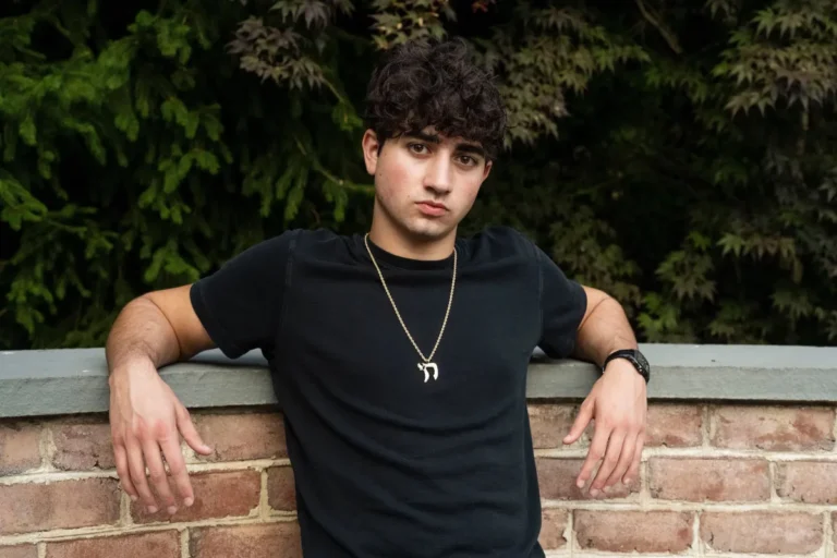 Person in a black shirt leans against a brick wall with greenery in the background, wearing a necklace and watch.