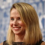 Woman with blonde hair smiling against a blue background with indistinct white text.