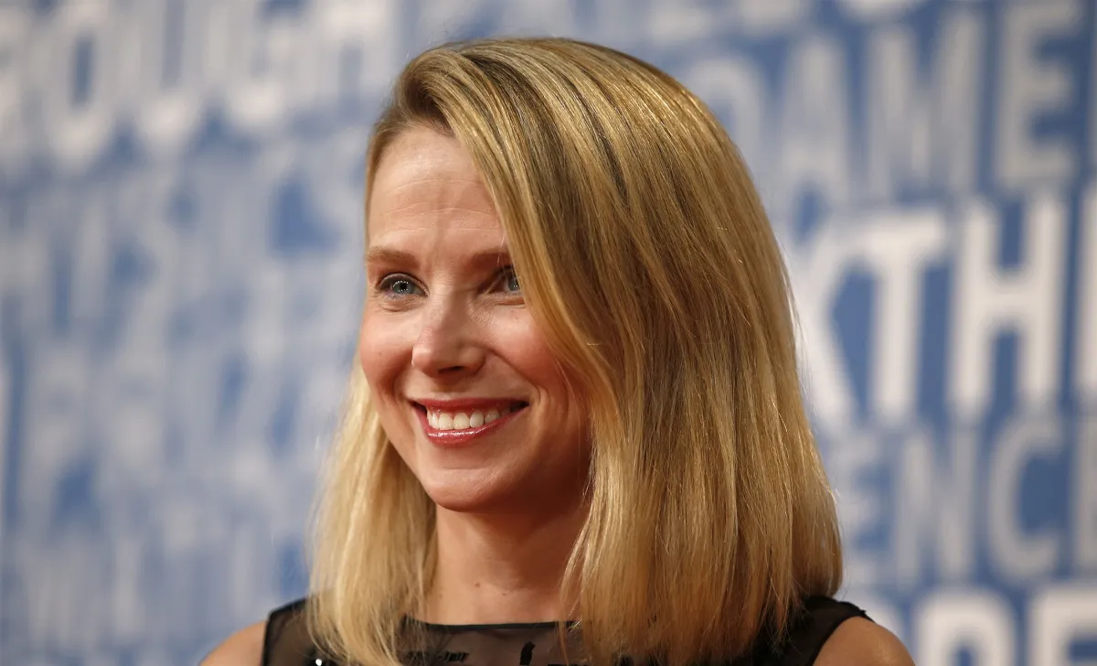 Woman with blonde hair smiling against a blue background with indistinct white text.