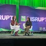 Two speakers sit on stage at TC Disrupt, green backdrop with "AI Stage" text and graphics, engaged in discussion.