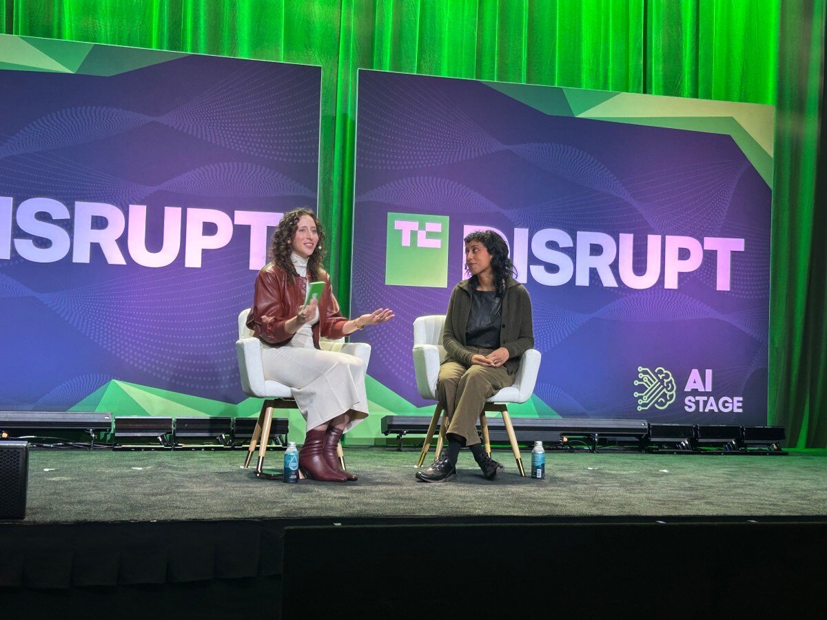 Two speakers sit on stage at TC Disrupt, green backdrop with "AI Stage" text and graphics, engaged in discussion.