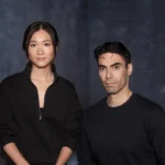 Two people in black clothing stand against a dark, textured backdrop, looking directly at the camera.