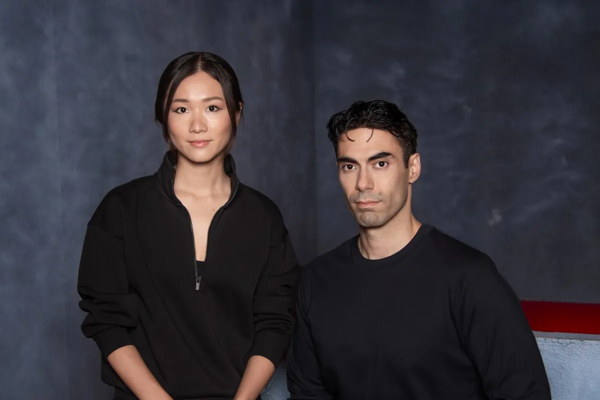 Two people in black clothing stand against a dark, textured backdrop, looking directly at the camera.