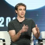Man in grey shirt speaking on a panel, seated in a white chair, gesturing with hands, blurred background with partial text.