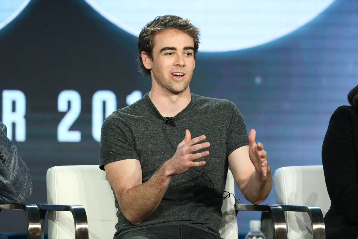 Man in grey shirt speaking on a panel, seated in a white chair, gesturing with hands, blurred background with partial text.