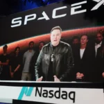 Large screen displays a person speaking at a podium with SpaceX logo, Nasdaq branding below, group of people in background.