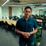 Man in Scimplify polo stands in modern office with long tables and chairs.