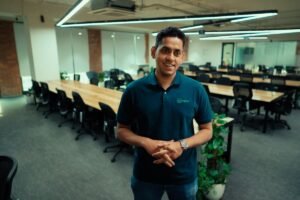 Man in Scimplify polo stands in modern office with long tables and chairs.