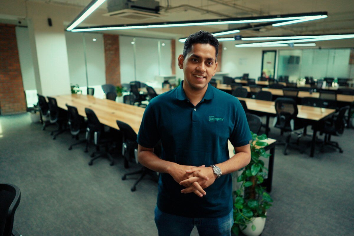 Man in Scimplify polo stands in modern office with long tables and chairs.
