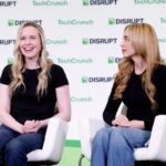 Two women seated on stage in front of TechCrunch Disrupt backdrop, both wearing black tops, one smiling, the other looking...