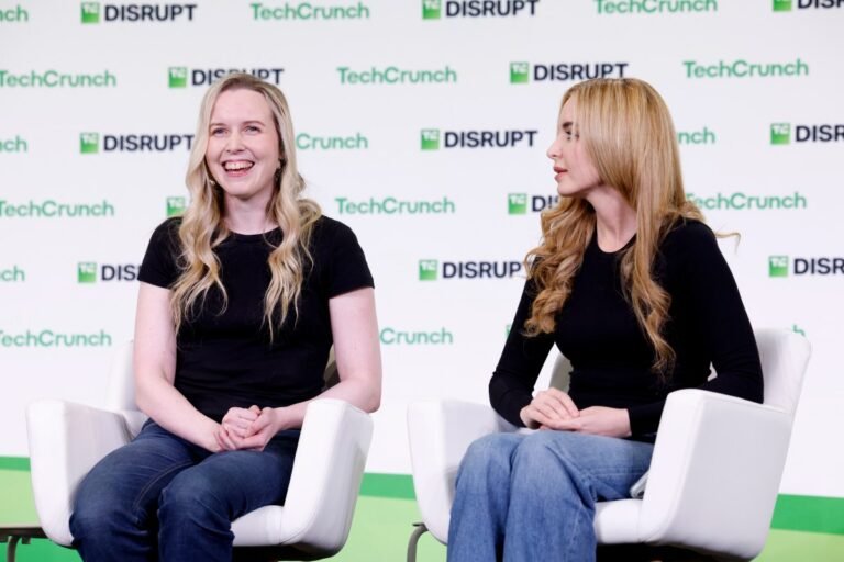 Two women seated on stage in front of TechCrunch Disrupt backdrop, both wearing black tops, one smiling, the other looking...